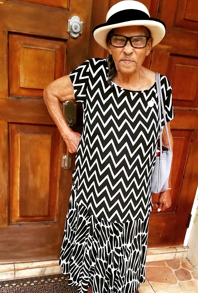 Leonie “Mama Lee” Clarke standing in her white hat and black-and-white dress at the wooden front doors of the family home.